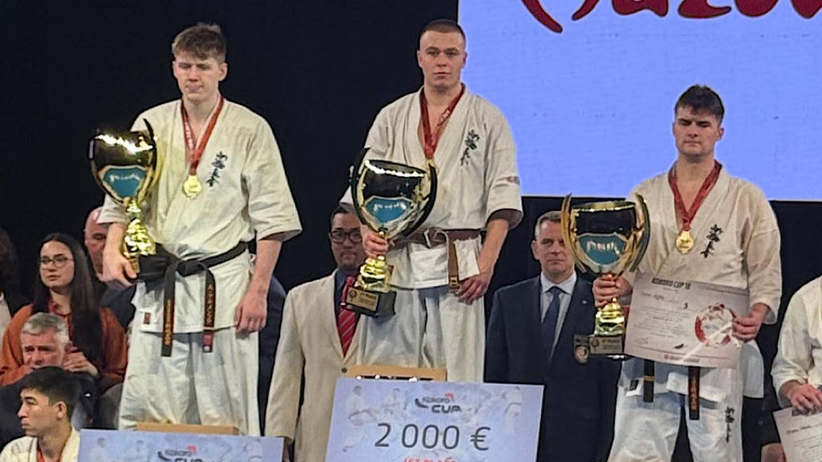 Trzech mężczyzn w strojach karate stoi na podium. Pierwszy od lewej ma na szyi złoty medal i trzyma puchar. Drugi od lewej, najwyższy, również ma medal i puchar. Trzeci od lewej ma medal i dokument. Na podium widoczne są tabliczki z kwotami 1000 €, 2000 € i 500 €.