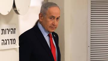 Portret polityka w garniturze i czerwonym krawacie, stojącego w pomieszczeniu.