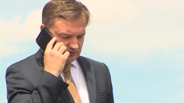 Mężczyzna w ciemnym garniturze i jasnej koszuli, z krawatem, rozmawia przez czarny smartfon trzymany przy uchu. Tło jest jasnoniebieskie.