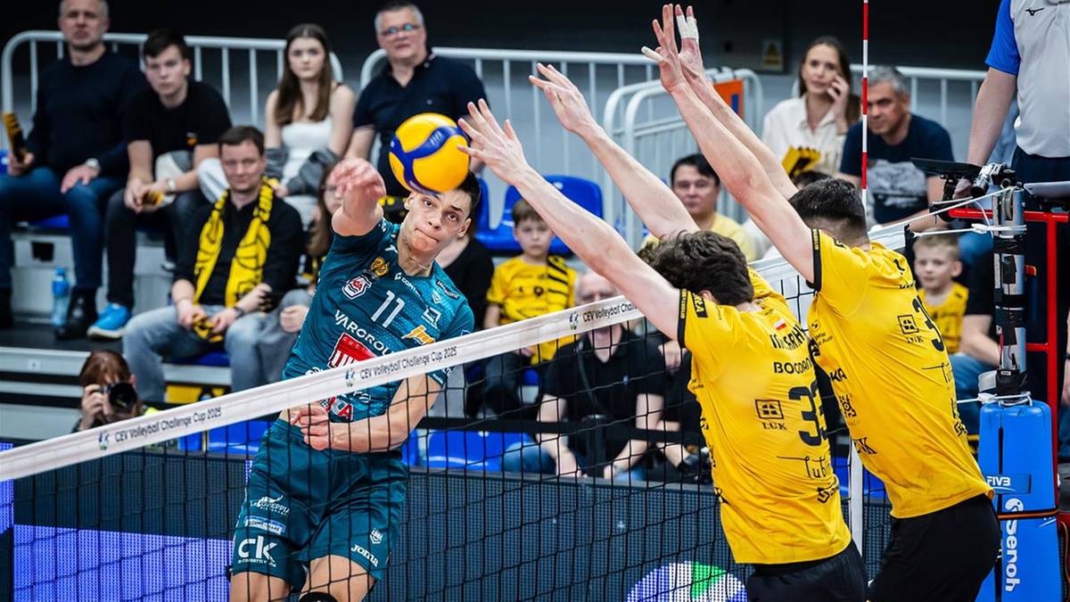 Siatkarz w niebieskim stroju atakuje piłkę nad siatką, blokowany przez dwóch przeciwników w żółtych strojach.