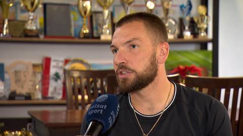 Portret Łukasza Kubota, tenisisty z kr&oacute;tką brodą i blond włosami, patrzącego w bok. Jest ubrany w czarną koszulkę z naszyjnikiem. W tle widać regały z trofeami i zdjęciami.