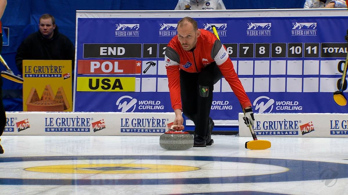 Zawodnik curlingu w czerwonej kurtce i czarnych spodniach wypuszcza kamień na lodowisku. W tle tablica wynik&oacute;w i logo "World Curling".