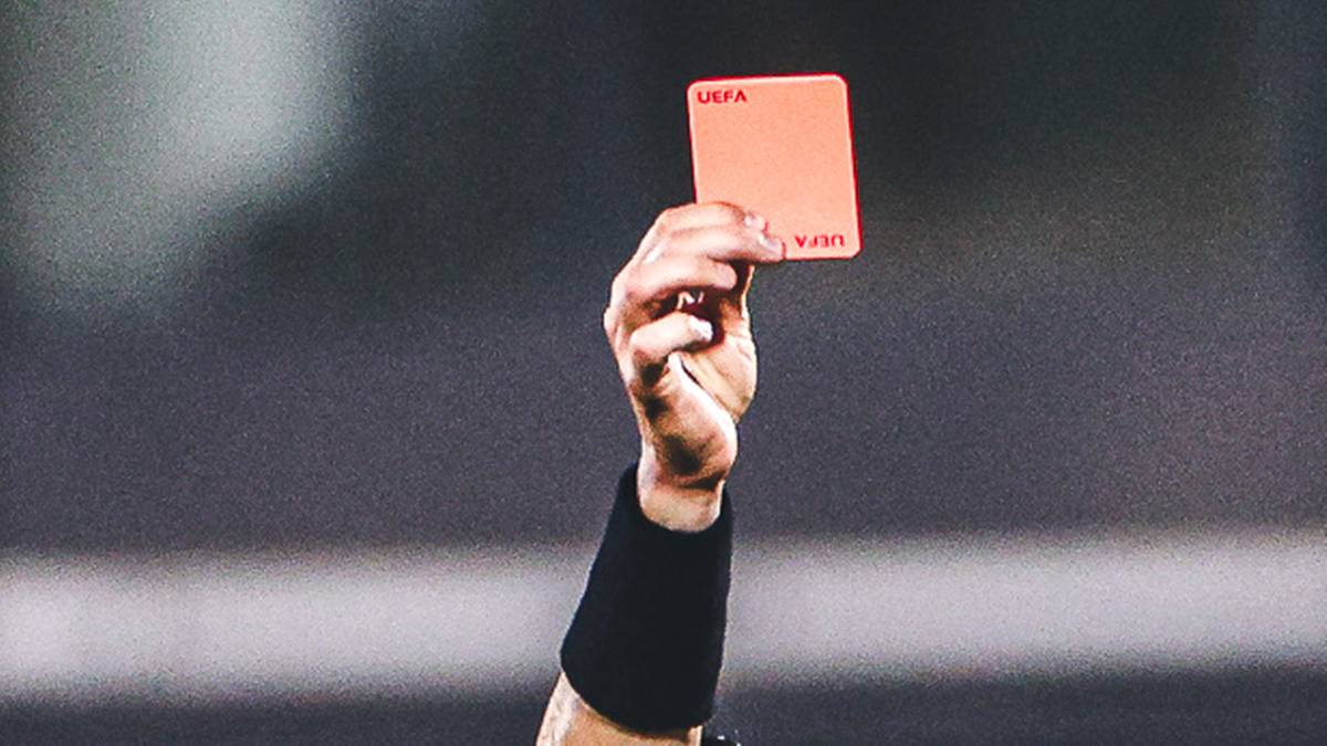 Sędzia piłkarski pokazuje czerwoną kartkę zawodnikowi, kt&oacute;ry jest pocieszany przez kolegę z drużyny.