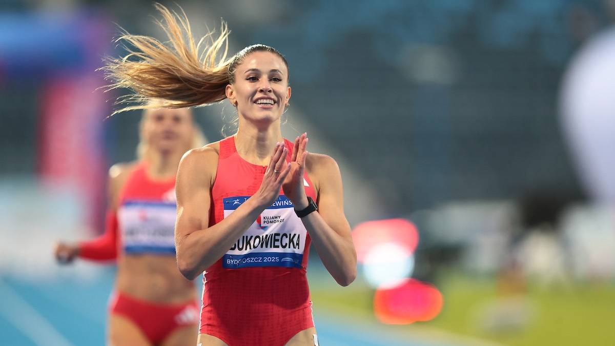 Atrakcyjna sportsmenka z blond włosami w czerwonym stroju lekkoatletycznym, biegnie i klaszcze w dłonie z uśmiechem na twarzy.