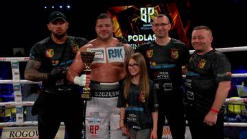 Pięciu mężczyzn i jedna dziewczynka stoją na ringu bokserskim, trzymając puchary i pozując do zdjęcia. Dw&oacute;ch mężczyzn po bokach ma na sobie czarne koszulki z logotypami, a mężczyzna pośrodku ma na piersiach napis "PJK". Dziewczynka stoi przed nim, a po jej lewej stronie znajduje się mężczyzna w okularach.