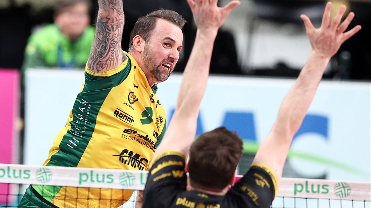 Siatkarz w żółtej koszulce z zielonymi pasami, z tatuażami na rękach, wykonuje akcję na siatce, podczas meczu siatkówki.