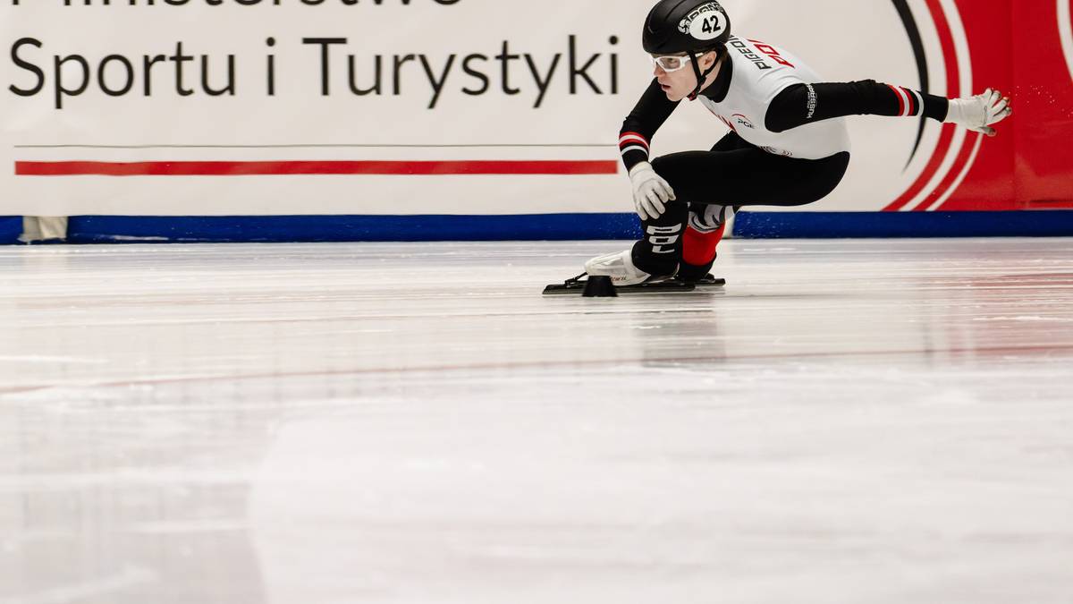 Zawodnik short track w stroju sportowym klęczy na lodzie, z kaskiem na głowie i rękami na śniegu. W tle znajduje się baner z logo Ministerstwa Sportu i Turystyki.