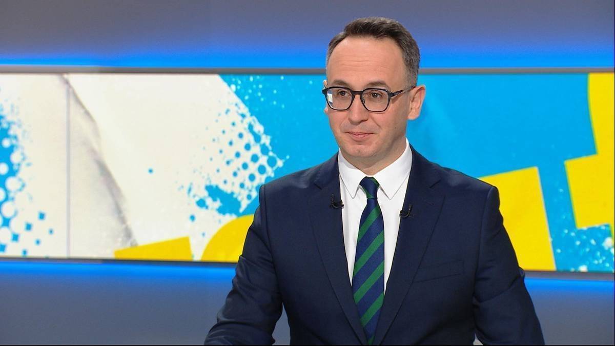 Mężczyzna w okularach i garniturze z krawatem stoi przed ekranem.