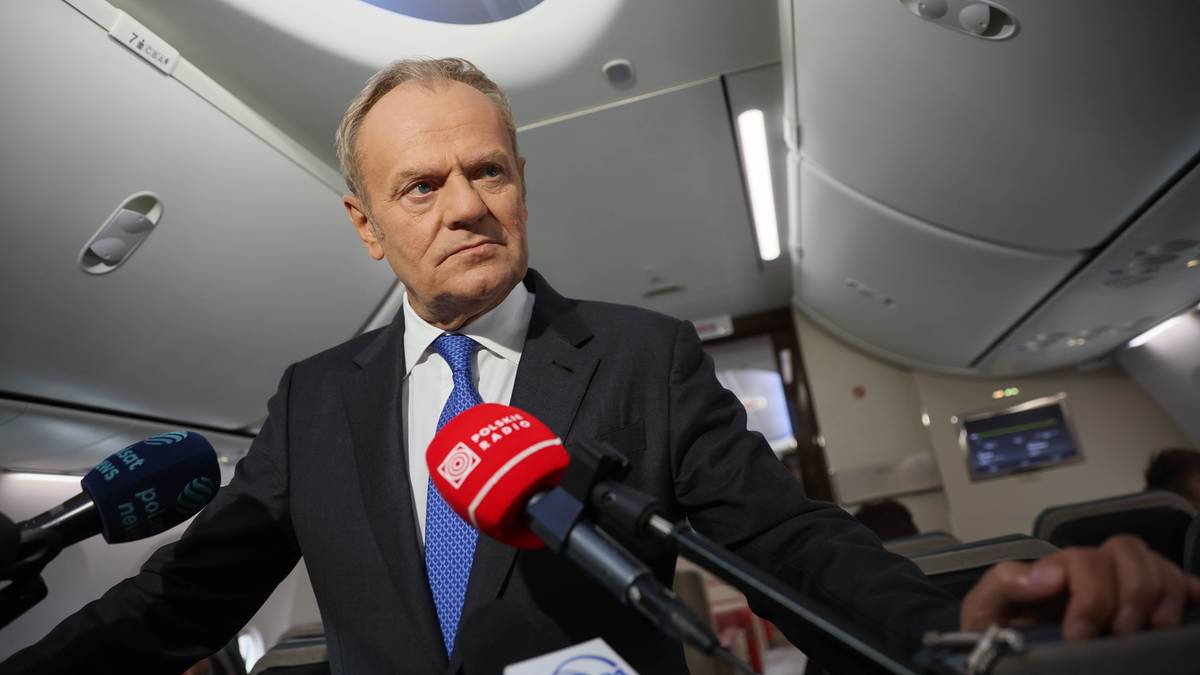 Polityk w garniturze i krawacie stoi przed mikrofonami wewnątrz samolotu.