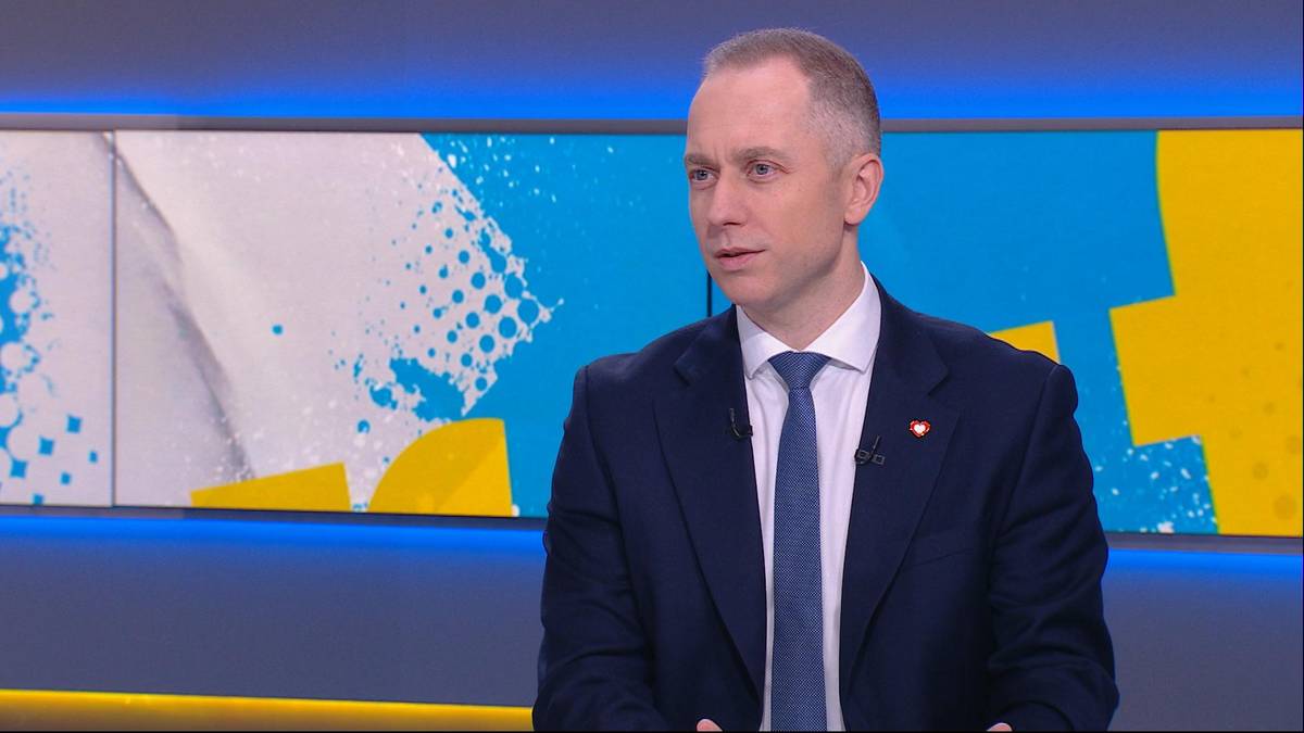 Mężczyzna w ciemnym garniturze i niebieskim krawacie siedzi w studiu telewizyjnym.