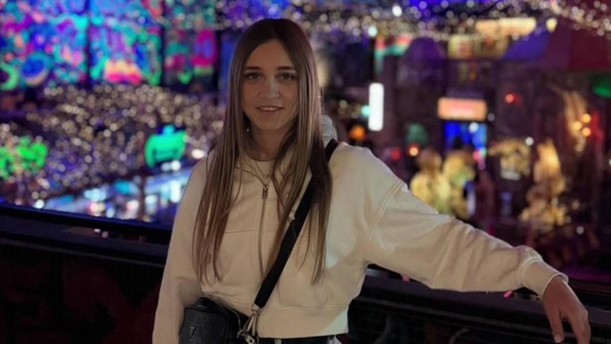 Młoda kobieta z długimi blond włosami uśmiecha się do aparatu. Jest ubrana w białą bluzę z kapturem i czarną torbę przewieszoną przez ramię. W tle widać kolorowe światła miasta nocą.