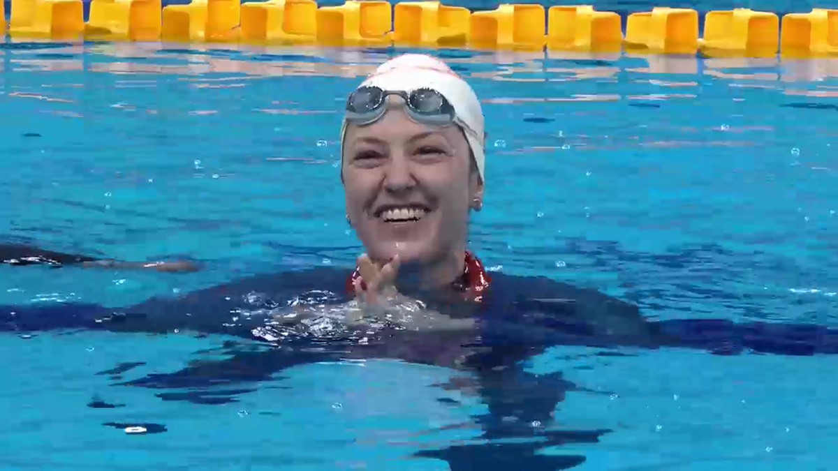 Uśmiechnięta pływaczka w czepku i okularach wychyla się z wody, z rękami złożonymi na piersi.