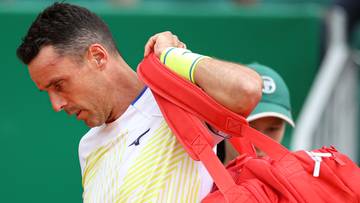 Tenisista z czerwoną torbą sportową na ramieniu, w białej koszulce z żółtymi pasami.