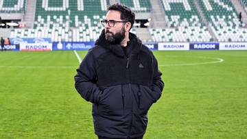 Mężczyzna w kurtce stoi na boisku piłkarskim przed trybunami stadionu z napisem Radomiak.