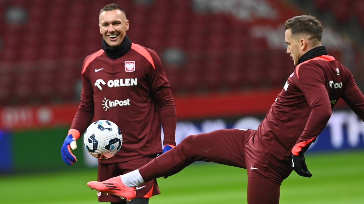 Dwóch polskich piłkarzy w bordowych strojach treningowych na boisku piłkarskim. Jeden z nich, stojący, trzyma piłkę pod lewą ręką i uśmiecha się. Drugi, w ruchu, kopie piłkę prawą nogą.