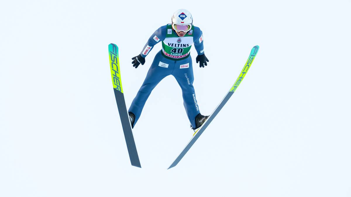 Skoczek narciarski w locie, w niebieskim kombinezonie, z nartami ustawionymi do lądowania.