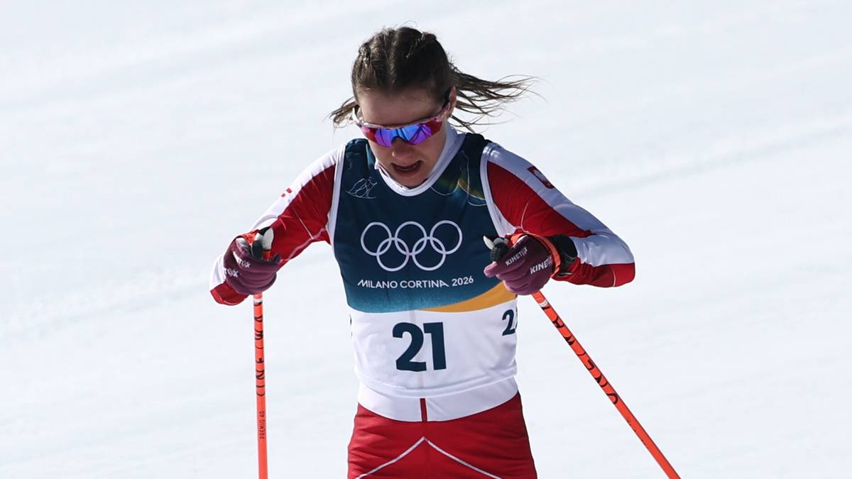 Kobieta w czerwonym stroju narciarskim i białej koszulce z numerem 21 na nartach biegowych, z kijkami w dłoniach, na ośnieżonym stoku z niebieskimi znacznikami trasy.