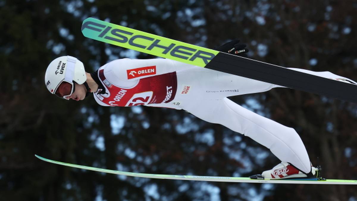 Skoczek narciarski w białym kombinezonie z czerwonymi elementami leci w powietrzu na nartach z nartami Fischer.