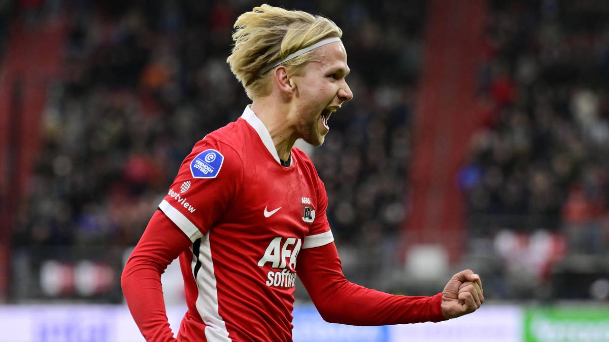 Piłkarz w czerwonej koszulce z blond włosami i opaską na czole krzyczy z radości, zaciskając pięść. Jest na boisku, podczas meczu.