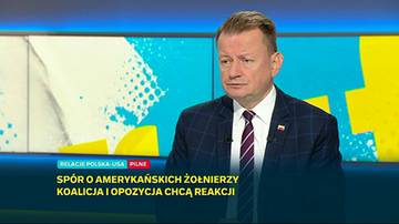 Mężczyzna w garniturze patrzy w bok, stojąc przed ekranem telewizyjnym z kolorowymi grafikami i napisem "RELACJE POLSKA-USA PILNE SP&Oacute;R O AMERYKAŃSKICH ŻOŁNIERZY KOALICJA I OPOZYCJA CHCĄ REAKCJI".