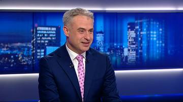 Mężczyzna w garniturze i krawacie w studiu telewizyjnym na tle panoramy miasta.