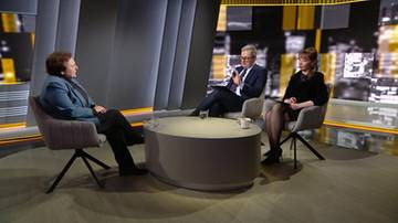 Trzy osoby siedzą w studiu telewizyjnym wok&oacute;ł okrągłego stołu. Po lewej siedzi kobieta w niebieskiej marynarce, zwr&oacute;cona twarzą do dw&oacute;ch pozostałych os&oacute;b. Pośrodku siedzi mężczyzna w garniturze i okularach, m&oacute;wiący z uniesioną dłonią. Po prawej siedzi kobieta w czarnej bluzce, słuchająca rozmowy.