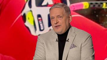 Portret Krzysztofa Rawy w jasnej marynarce i czarnym golfie, na tle czerwonego elementu sportowego.