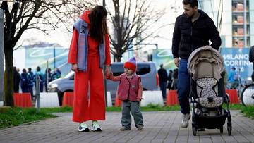 Kobieta w czerwonym dresie i mężczyzna w czarnej kurtce spacerują z małym dzieckiem i w&oacute;zkiem dziecięcym po chodniku.