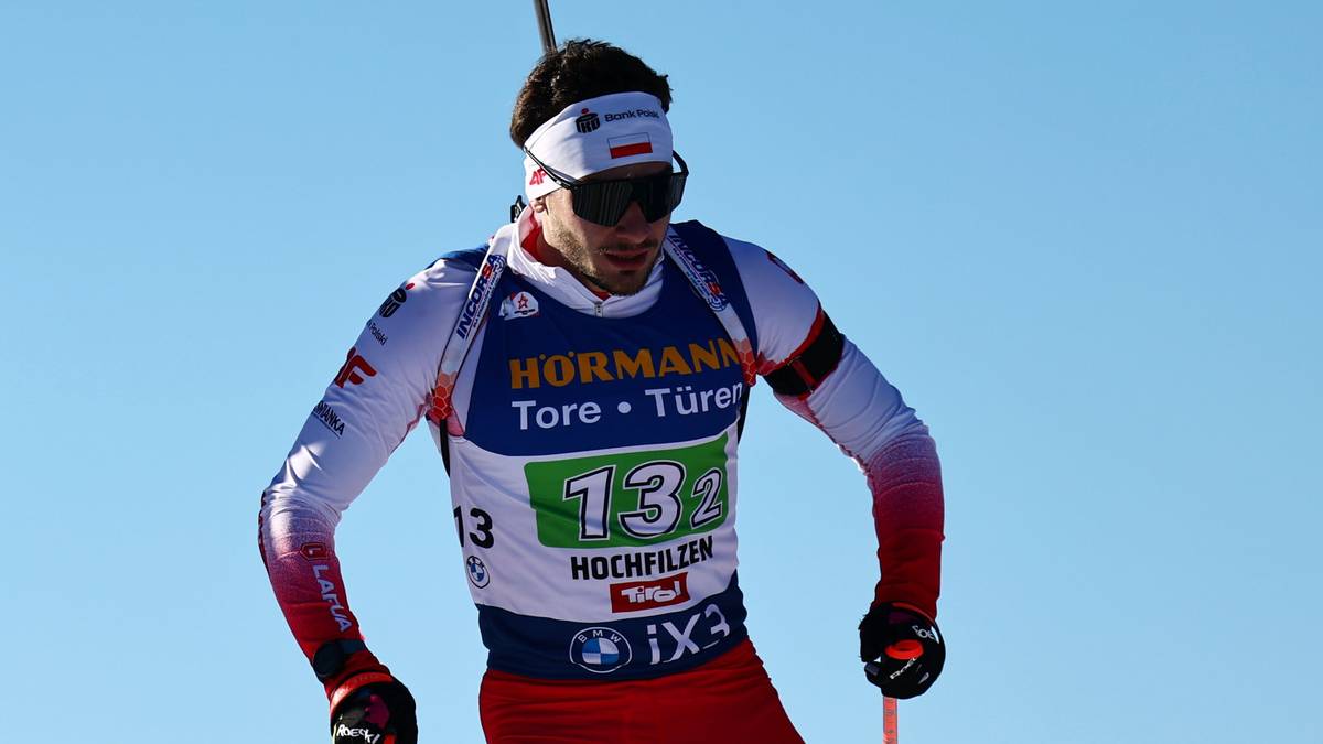 Dw&oacute;ch biathlonist&oacute;w w akcji na śniegu, jeden w niebieskim stroju z numerem 16, drugi w białym i czerwonym z numerem 13, obaj trzymający kije narciarskie.