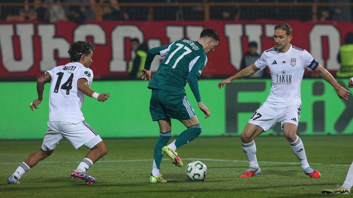 Piłkarze w białych i zielonych strojach rywalizują o piłkę na boisku.