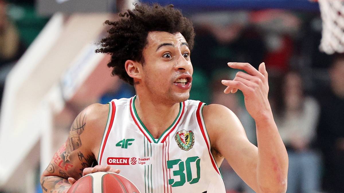 Koszykarz w białej koszulce z numerem 30 i zielonymi wstawkami, trzymający pomarańczową piłkę do koszyk&oacute;wki i gestykulujący ręką.