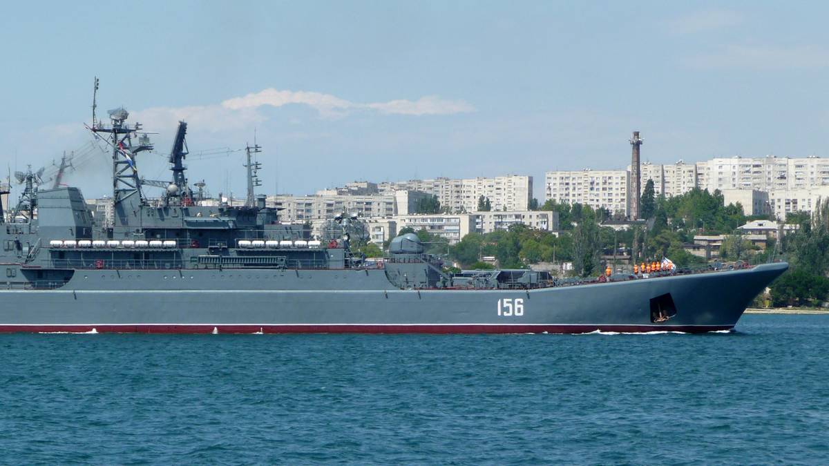 Okręt wojenny z numerem burtowym 156 płynie po wodzie na tle zabudowy mieszkalnej i budowli przemysłowych.