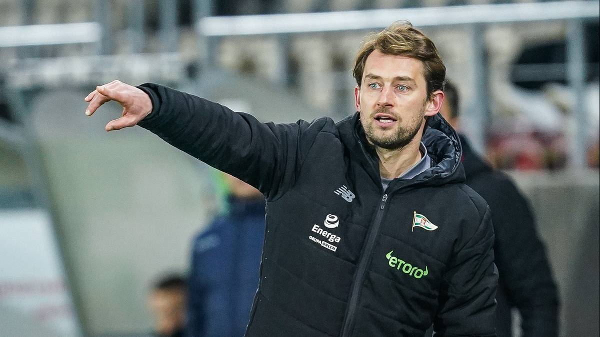 Trener Kaczmarek w czarnej kurtce z napisem "TK" i czerwonymi akcentami na butach, wskazujący ręką na boisku piłkarskim.