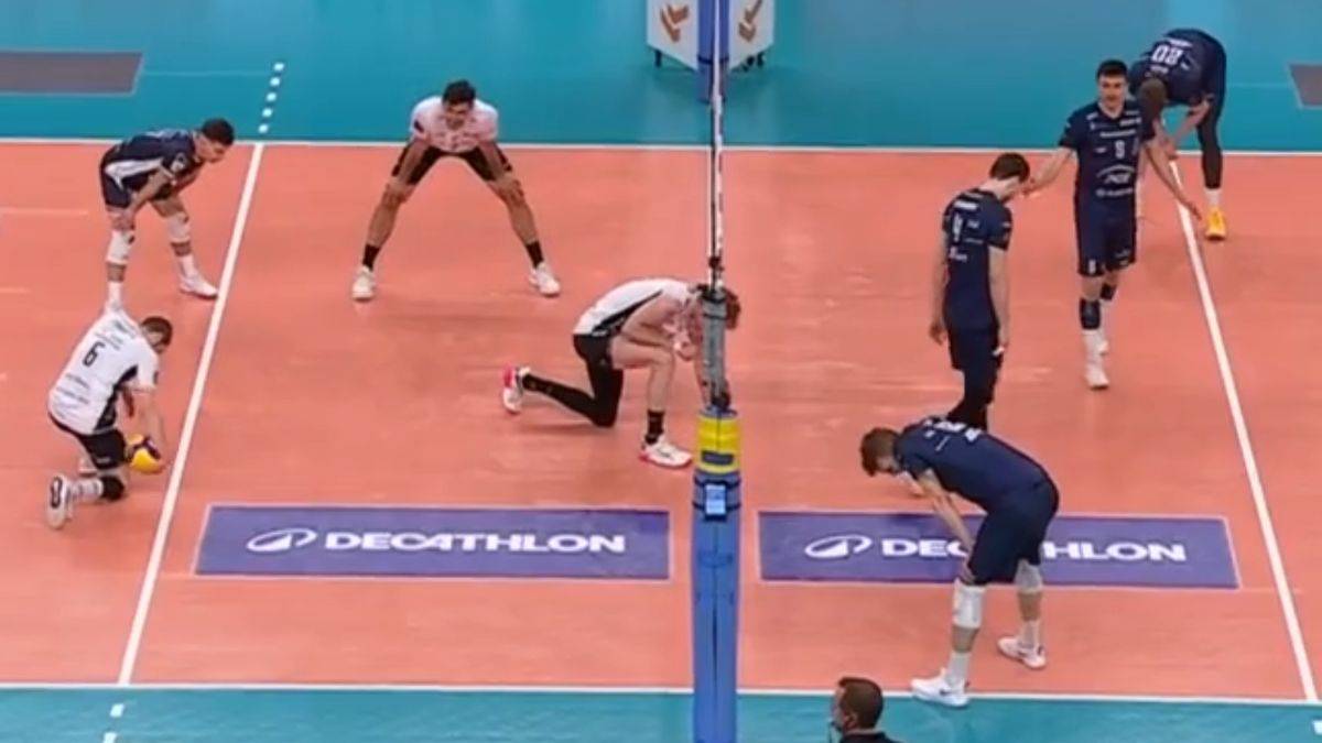 Sześciu graczy w niebieskich i białych strojach gra w siatkówkę na boisku z pomarańczową nawierzchnią. Dwóch graczy pochyla się, żeby odebrać piłkę, podczas gdy inni stoją w gotowości. W tle widać niebieską siatkę.