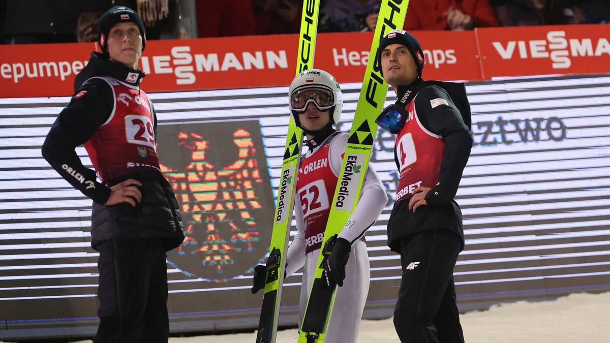 Trzech skoczków narciarskich w strojach sportowych pozuje do zdjęcia, trzymając narty.