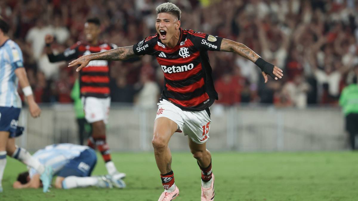 Piłkarz w czarno-czerwonej koszulce z białymi spodenkami, z uniesionymi rękami i krzykiem radości, biegnie po zielonym boisku piłkarskim. W tle widać innych piłkarzy i rozmazanych kibiców na trybunach.