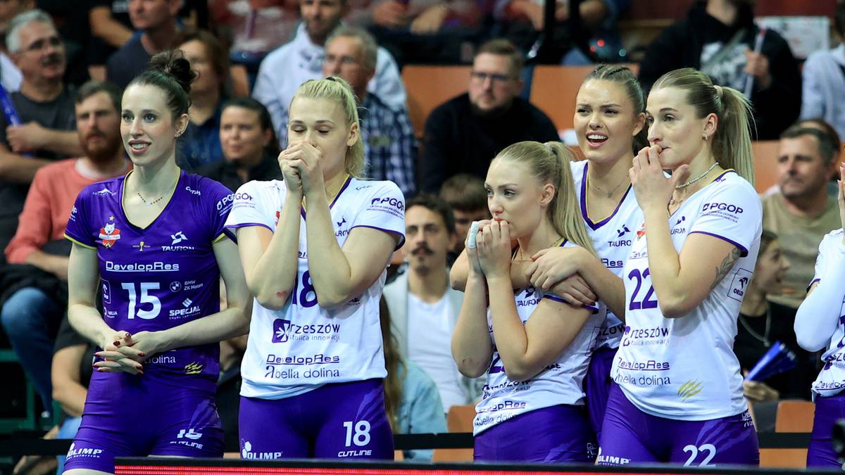 Grupa siatkarek w białych strojach sportowych trzyma ręce przy ustach.