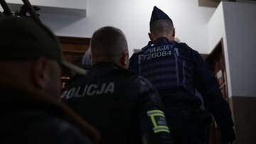 Dw&oacute;ch policjant&oacute;w w czarnych mundurach z napisem POLICJA na plecach, widzianych od tyłu w budynku.
