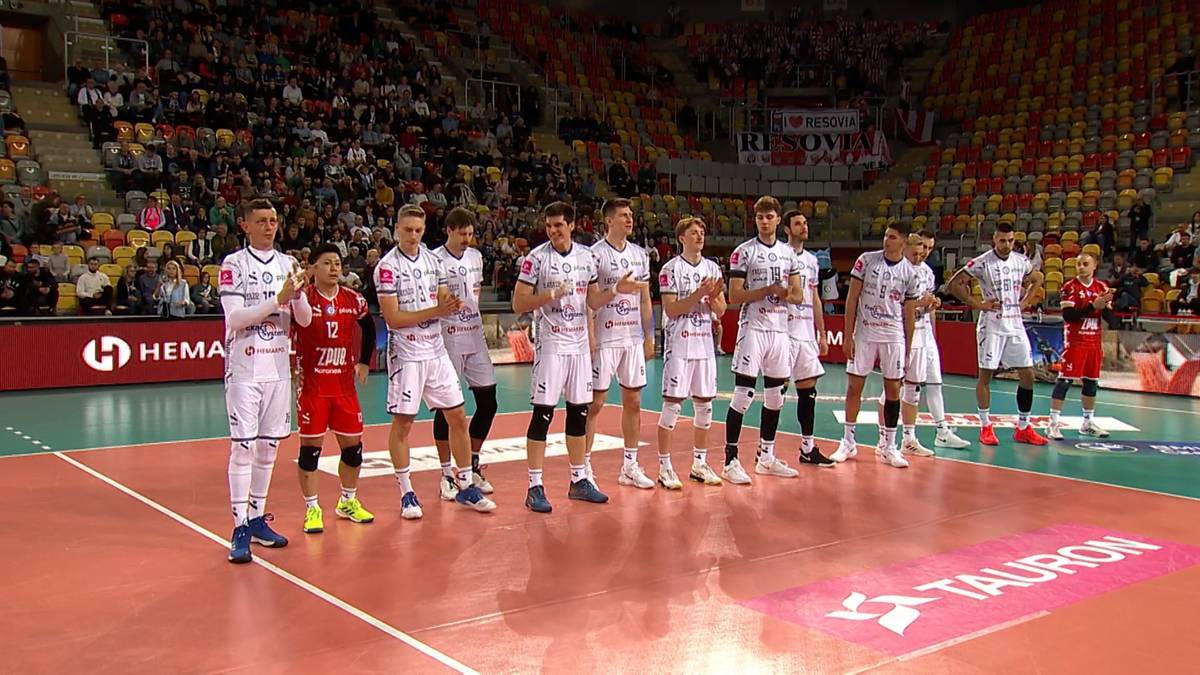 Drużyna siatkarzy w białych strojach stoi na parkiecie hali sportowej, zwr&oacute;cona w stronę widowni.