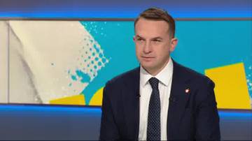 Adam Szłapka siedzi w studiu telewizyjnym, ubrany w ciemny garnitur, białą koszulę i granatowy krawat w białe kropki. Tło stanowi niebieska ściana z abstrakcyjnymi ż&oacute;łtymi i białymi elementami graficznymi.