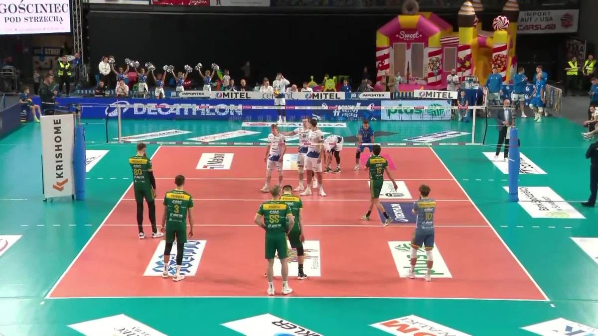 Boisko do siatkówki z zawodnikami w trakcie meczu, otoczone trybunami z publicznością.