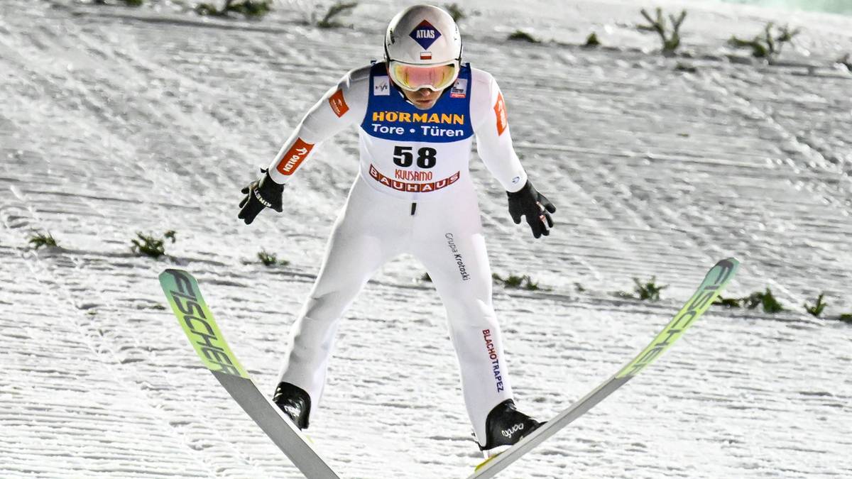 Skoczek narciarski w białym kombinezonie z numerem 58 i pomarańczowymi wstawkami, w białym kasku i goglach, stoi na skoczni narciarskiej z nartami ustawionymi na belce startowej.