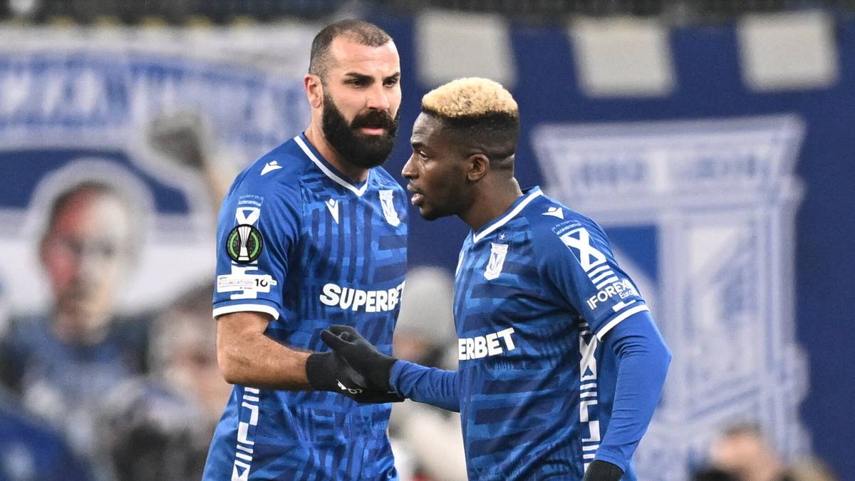 Dwóch piłkarzy Lecha Poznań w niebieskich strojach stoi na boisku, rozmawiając ze sobą i ściskając sobie dłonie.