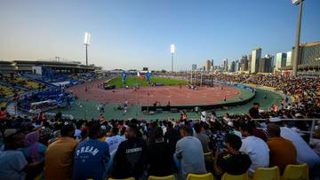 Widok na stadion lekkoatletyczny z trybunami wypełnionymi widzami i bieżnią z zawodnikami w tle panoramy miasta.