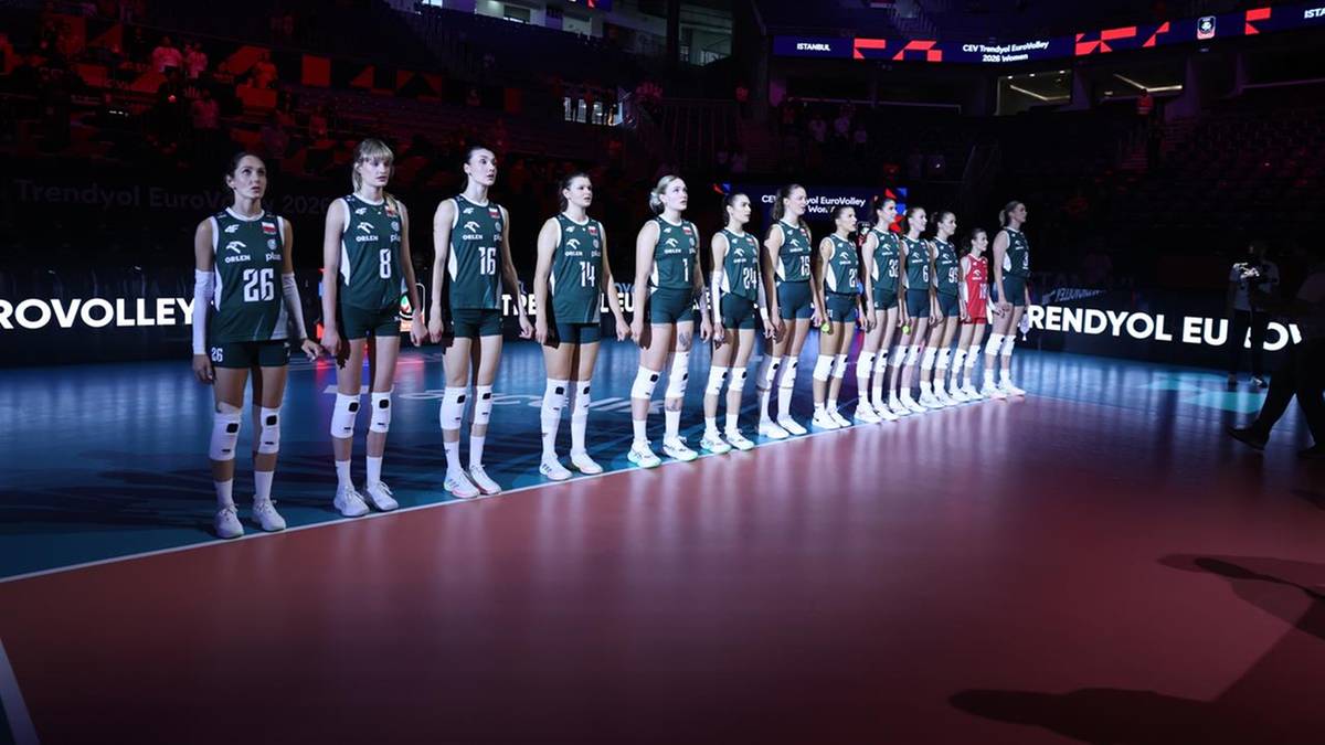 Drużyna siatkarek w strojach sportowych stoi w rzędzie na boisku, z widownią w tle.