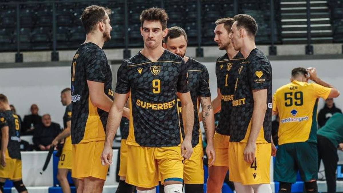 Siatkarze w ż&oacute;łto-czarnych strojach stoją na boisku hali sportowej.