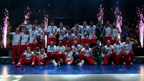 Drużyna polskich siatkarzy w białych strojach z czerwonymi elementami pozuje na podium z pucharem, w tle fajerwerki.
