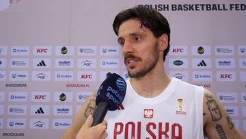 Dominik Olejniczak, koszykarz reprezentacji Polski, udziela wywiadu podczas wydarzenia sportowego. Na jego koszulce widnieje napis "POLSKA" i orzeł.