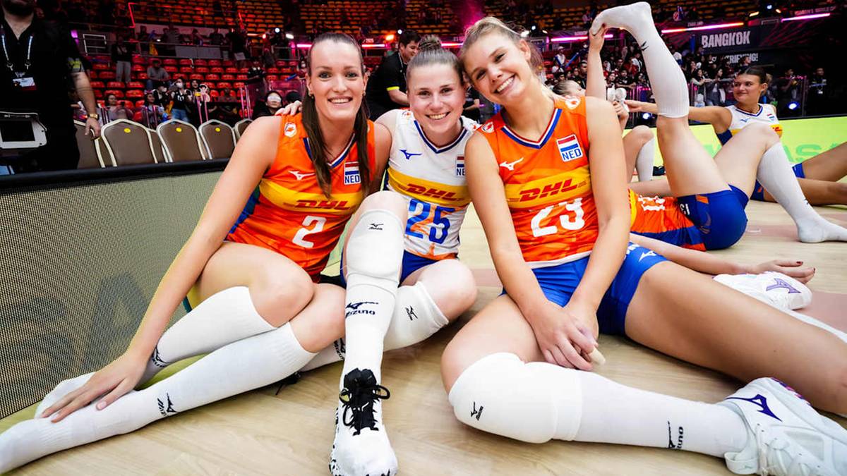 Trzy zawodniczki siatkówki w pomarańczowych strojach reprezentacji Holandii siedzą na podłodze, uśmiechając się do kamery po zwycięskim meczu.