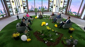 Siedem os&oacute;b siedzi w okręgu na zielonej wykładzinie w studiu telewizyjnym. W tle widać panoramę miasta.